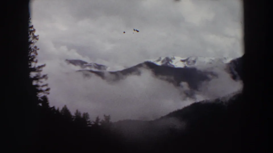 1964 Washington 8mm frame showing clouded snow-capped mountains beyond a dark forested foreground.