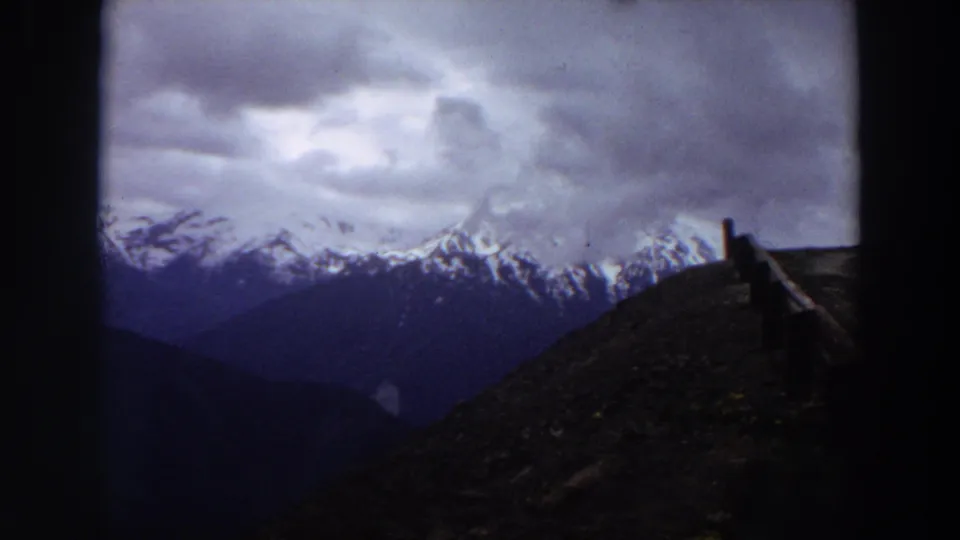 1964 Washington 8mm frame of snow-capped mountains beneath clouds, seen from a roadside with a wooden guardrail.
