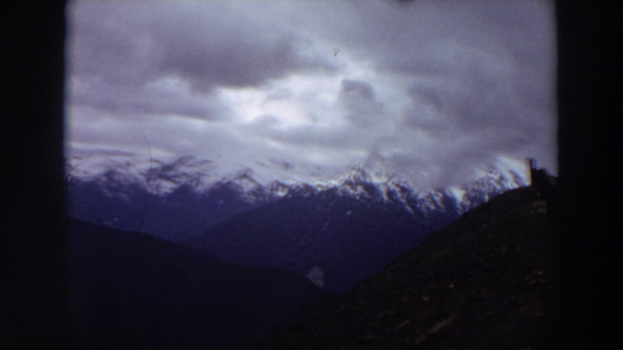 1964 Washington 8mm frame showing distant snow-capped mountains beneath a brooding cloudy sky, partly obscured by dark foreground edges.