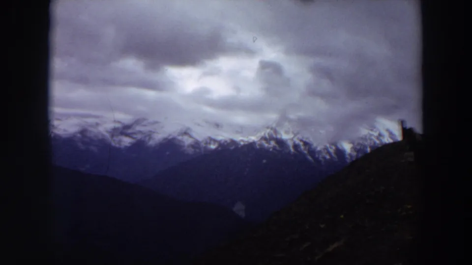 1964 Washington 8mm frame showing distant snow-capped mountains beneath a brooding cloudy sky, partly obscured by dark foreground edges.