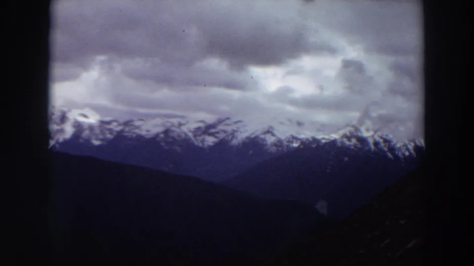 1964 Washington 8mm frame of a snowy mountain ridge and layered foothills beneath dense gray clouds, with dark film or window edges.