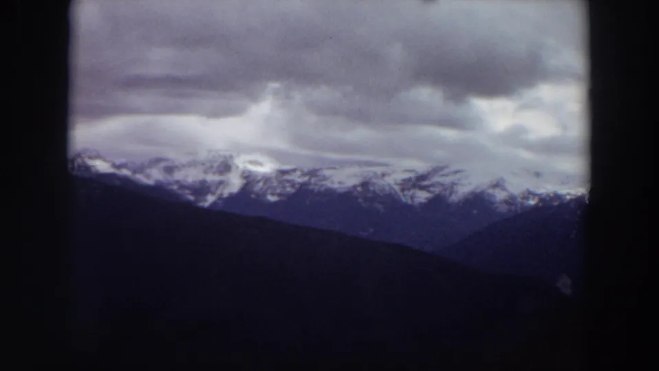 1964 Washington 8mm frame showing a distant snow-capped range, dark foothills, and shifting gray clouds across the upper frame.