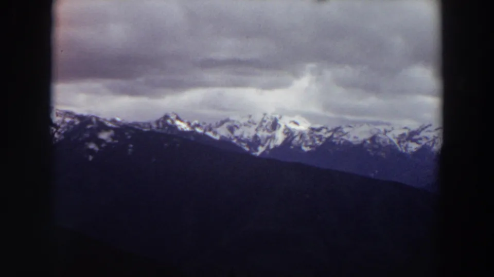 1964 Washington 8mm frame of snow-streaked mountain peaks beneath dense clouds, with dark foreground framing along the right side.
