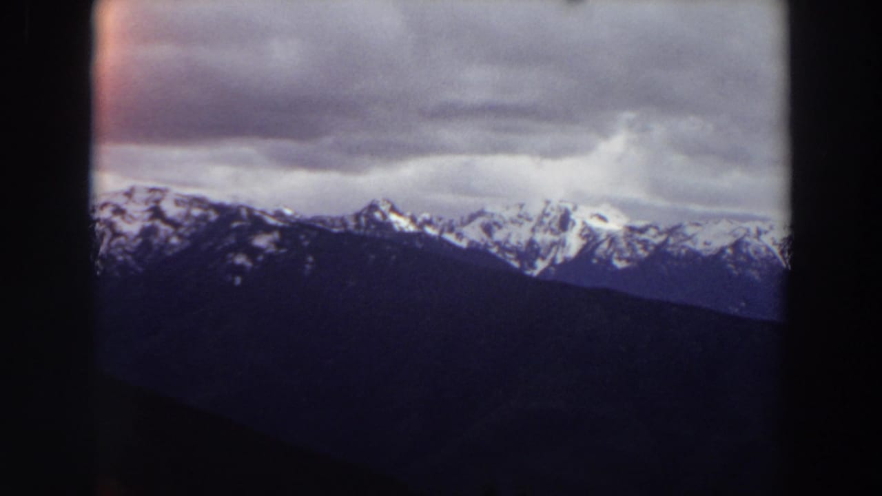 1964 Washington 8mm frame showing distant snow-covered peaks beneath low gray clouds, framed by dark projection edges.