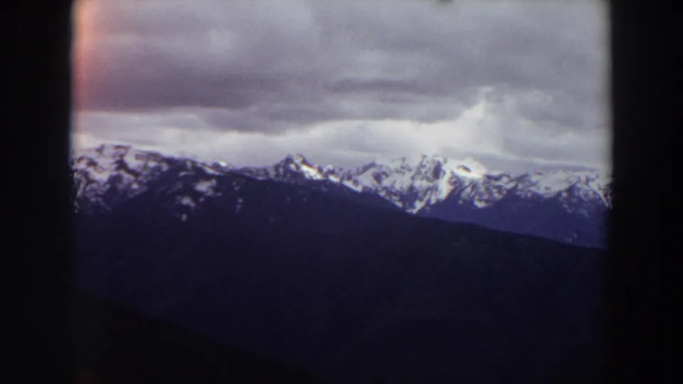 1964 Washington 8mm frame showing distant snow-covered peaks beneath low gray clouds, framed by dark projection edges.