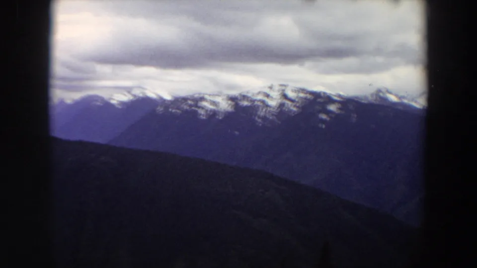 A soft 8mm view of Washington mountains shows dark foreground slopes, distant snowy ridgelines, and a thick gray cloud bank.