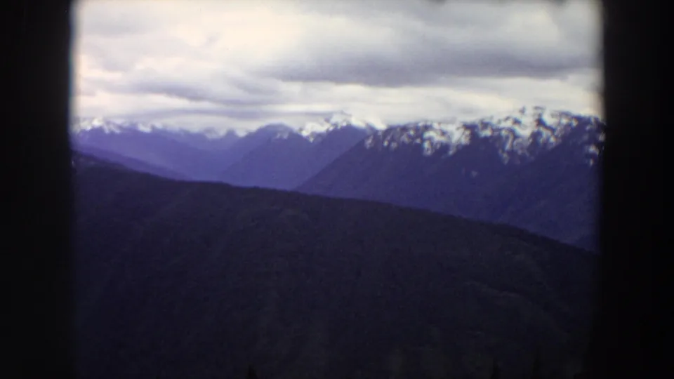 Washington mountain layers fill the frame, with snow on the far peaks, a cloudy sky above, and dark 8mm projection borders around the view.