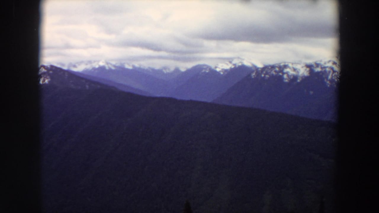 1964 Washington 8mm frame showing layered mountain ridges with snow patches under an overcast sky and dark projection borders.