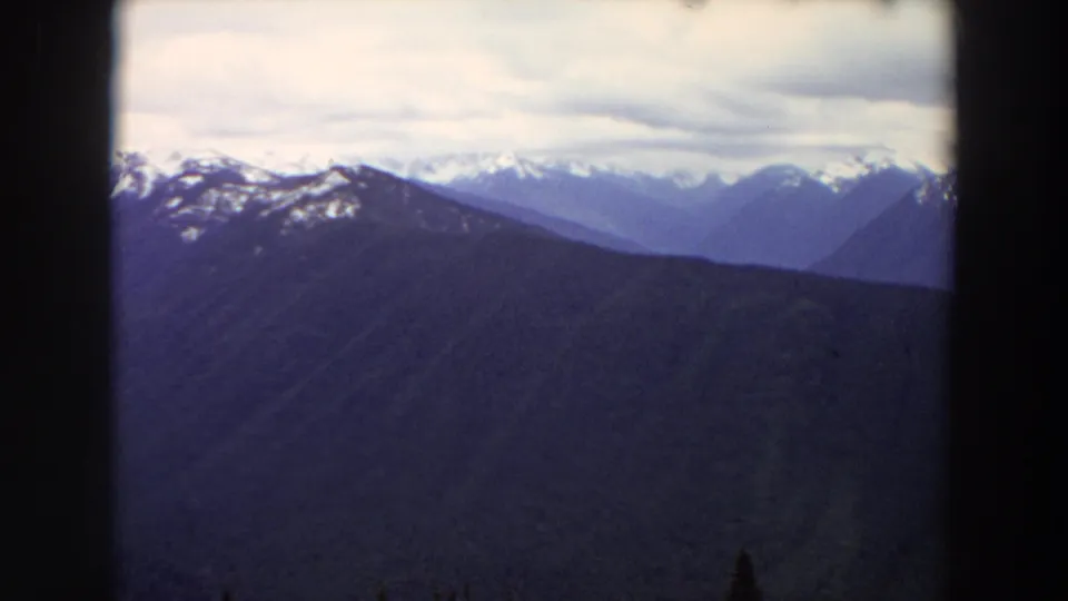 1964 Washington scenic frame with blue mountain layers, snow on distant ridges, overcast clouds, and visible 8mm frame edges.