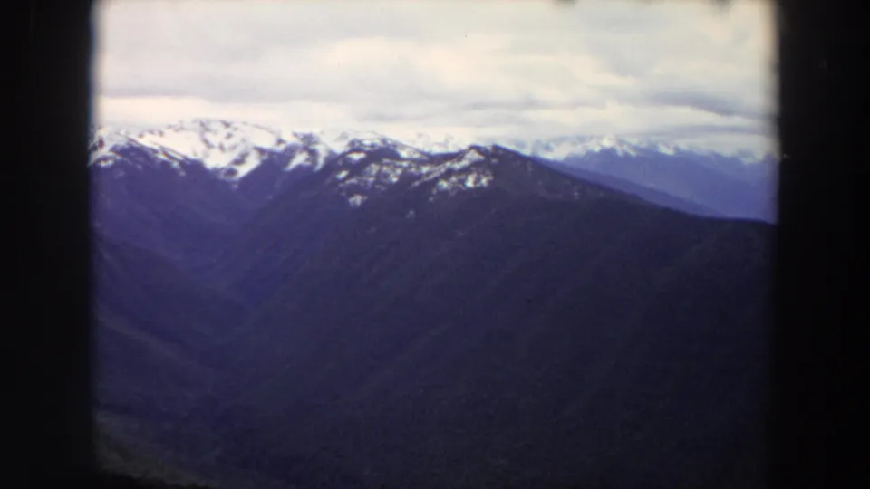 1964 Washington home-movie frame showing snow-streaked peaks above darker mountain slopes, with cloudy sky and 8mm softness.