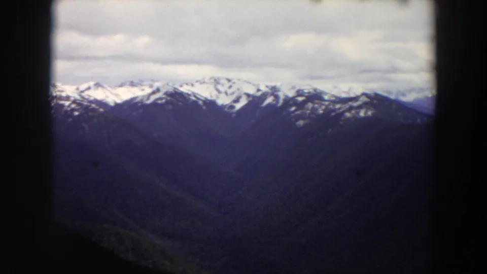 1964 Washington 8mm frame with a broad snow-streaked mountain range, dark lower ridges, cloudy sky, and visible projection borders.