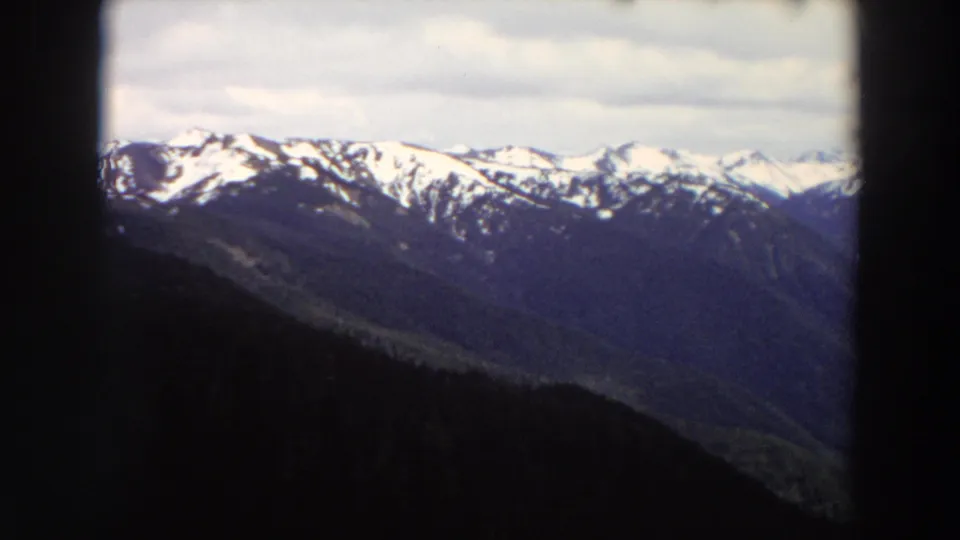 1964 Washington 8mm frame of a snow-streaked mountain ridge above a shadowed forest valley.