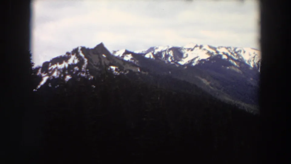 1964 Washington home-movie frame with jagged snow-covered peaks, forested slopes, and dark film-gate edges.