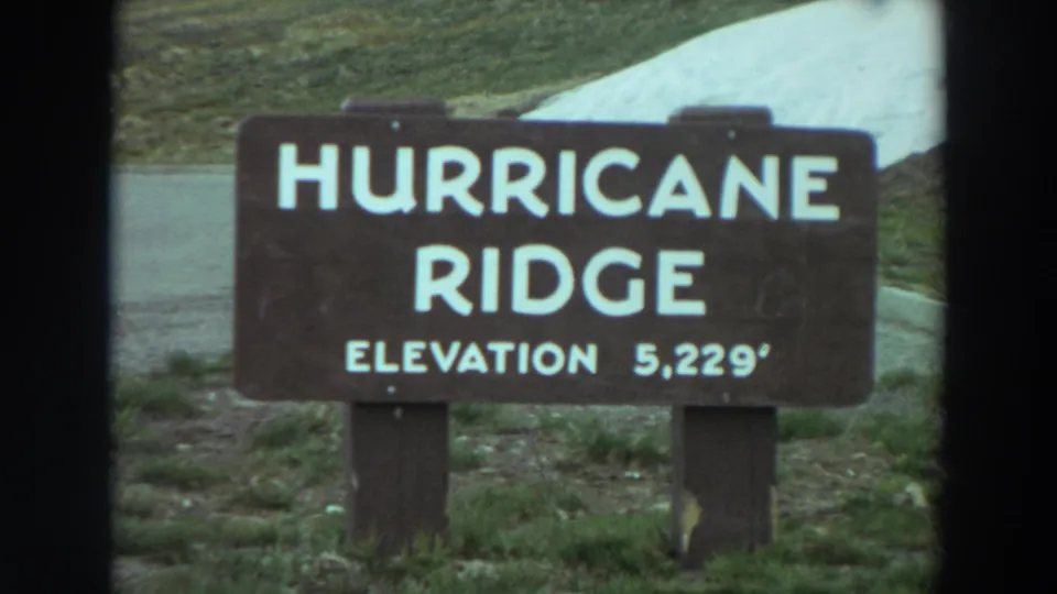 1964 Washington 8mm frame showing a brown Hurricane Ridge sign marked elevation 5,229 feet beside a roadside slope.