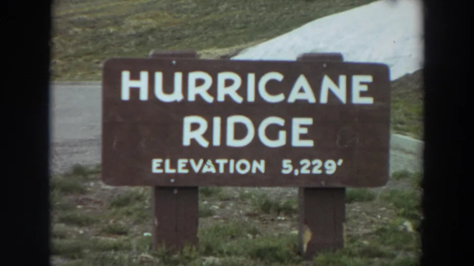 8mm home-movie frame of the Hurricane Ridge elevation sign, with grass, pavement, and snow-covered ground in the background.