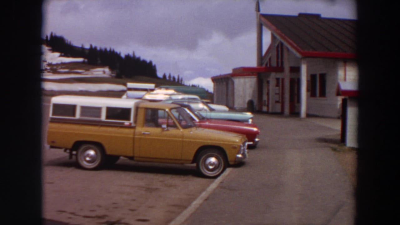 1964 Washington 8mm frame showing an orange pickup with camper shell parked near classic cars outside a white roadside building.
