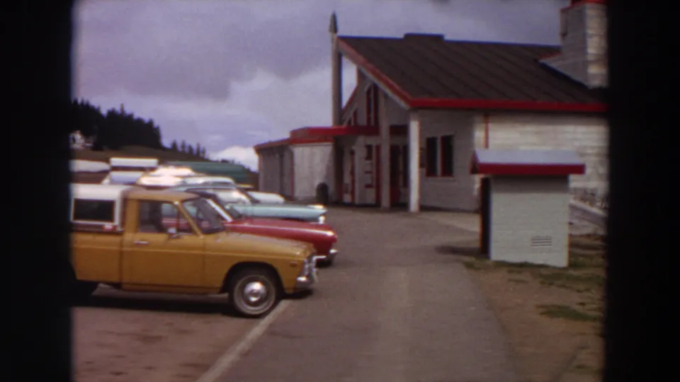 1964 Washington home-movie frame with a parked orange pickup, classic cars, and a white roadside building with red trim.