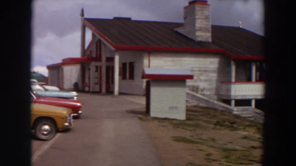 1964 Washington frame as the camera pans toward a white building with red trim, a stone chimney, and cars at the edge.