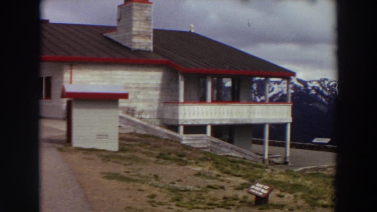 1964 Washington 8mm frame showing a stone-and-wood mountain lodge, balcony, path, and distant snow-capped peaks.