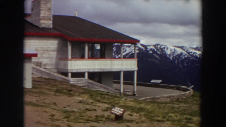 1964 Washington home-movie frame with a red-trimmed lodge, overlook sign, and snow-capped mountain range in the background.