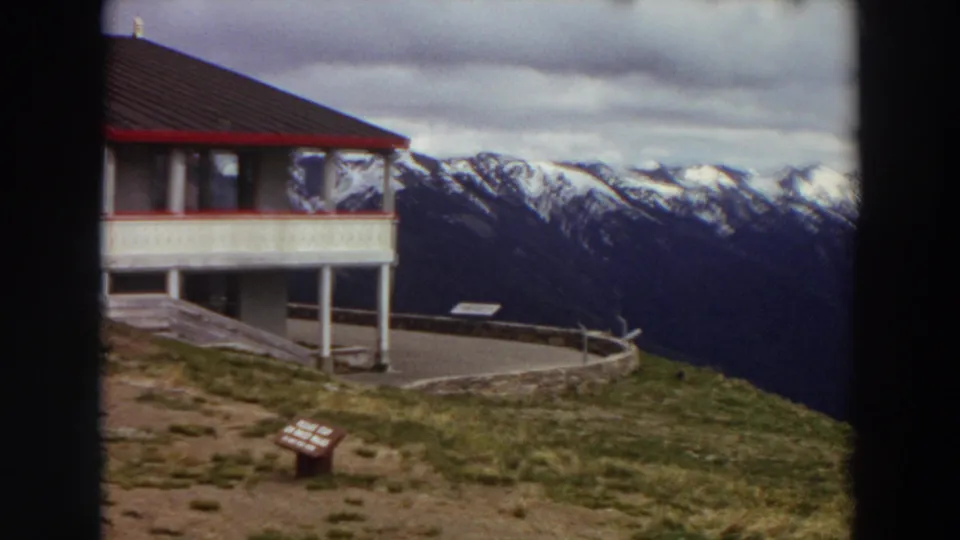 1964 Washington 8mm frame showing the edge of a mountain lodge, paved viewpoint, railing, and distant snowy peaks.
