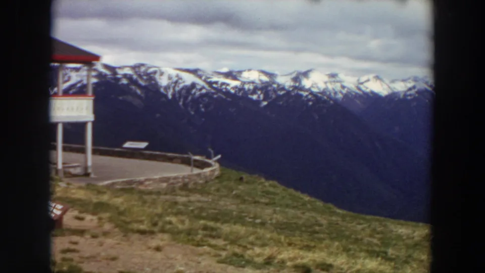 1964 Washington home-movie frame with the lodge edge, grassy overlook, small sign, and broad snow-capped peaks under clouds.