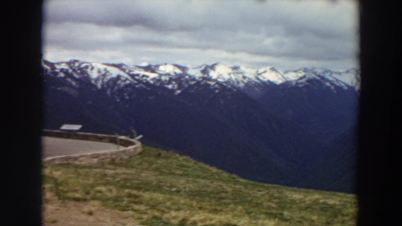 1964 Washington 8mm frame showing snowy mountain peaks, dark ridges, cloudy sky, and a grassy overlook with a small structure at left.