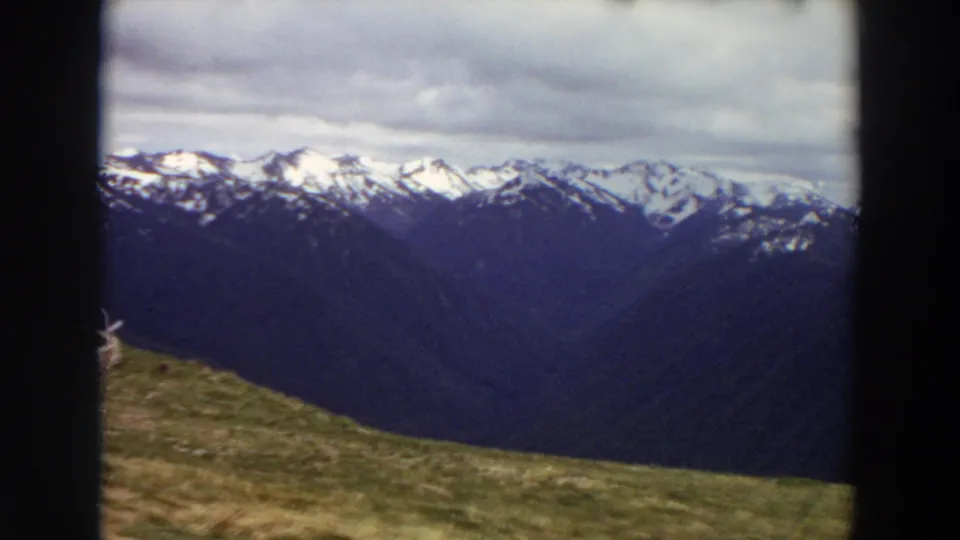1964 Washington 8mm view across a grassy foreground toward layered mountain ridges and snow-lined peaks beneath a cloudy sky.