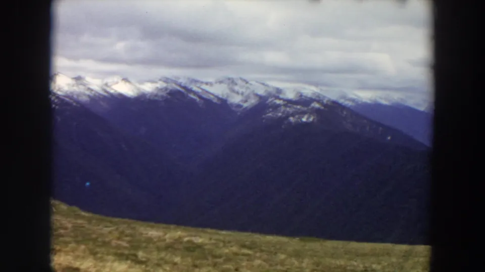 1964 Washington home-movie frame of blue-dark ridges, distant snow patches, heavy clouds, and a sloping grassy foreground.