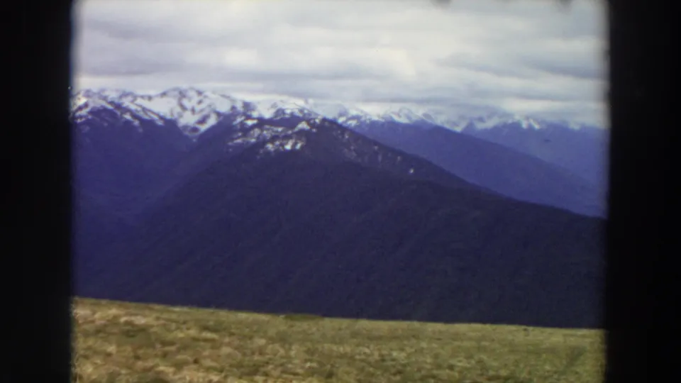 1964 Washington 8mm frame showing snow-dusted ridgelines, a cloudy sky, and a muted grassy foreground from a high overlook.
