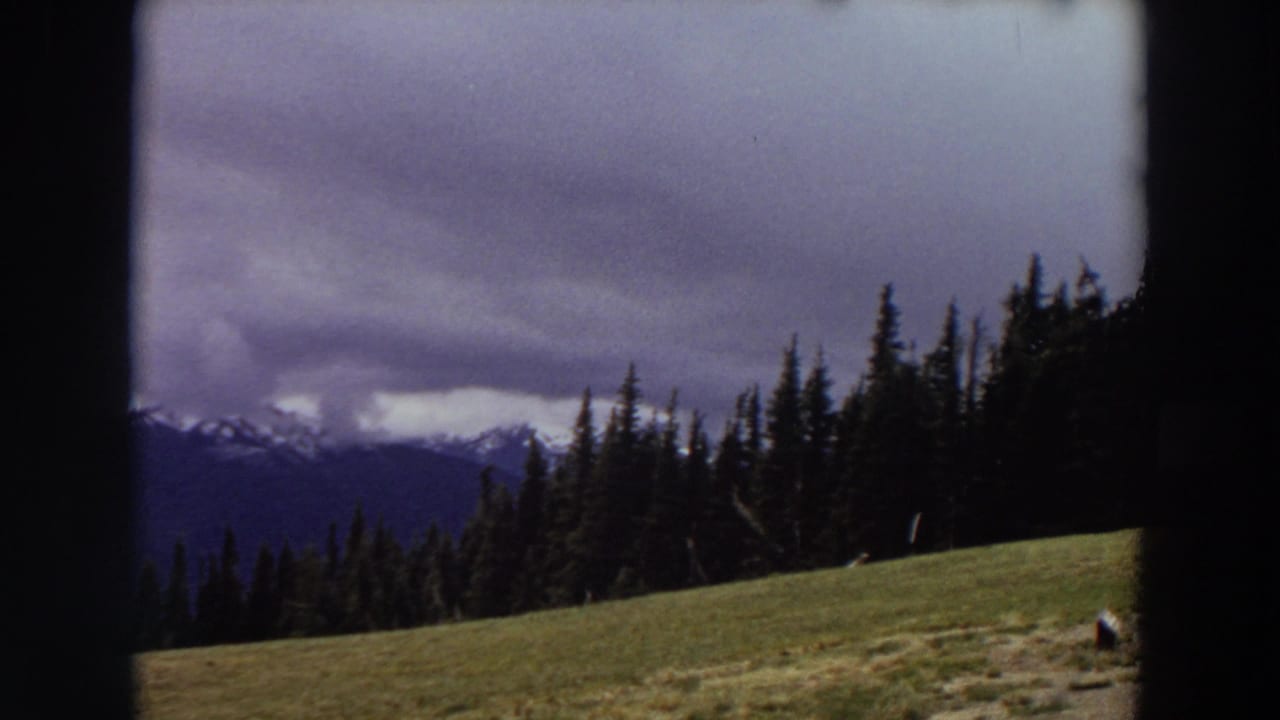 1964 Washington 8mm frame showing distant snow-capped mountains, evergreen forest, and a sloping meadow beneath heavy cloud cover.