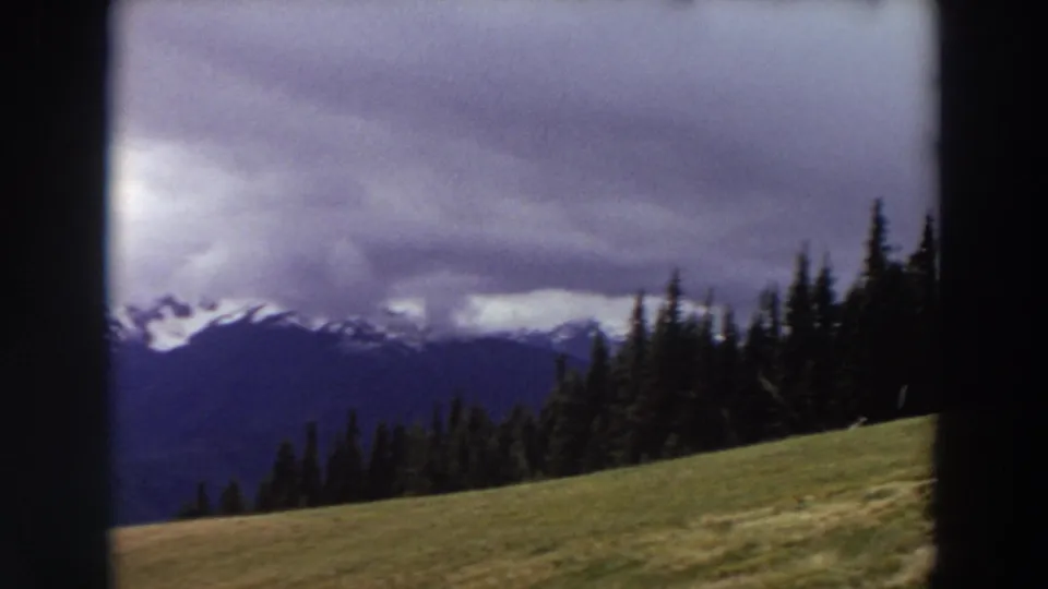 1964 Washington landscape frame with a forested ridge, open hillside, and distant snow-lined peaks beneath overcast sky.