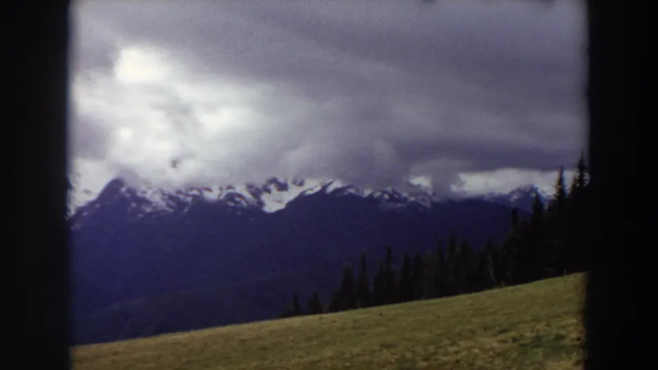 1964 Washington 8mm view of mountain peaks, evergreen forest, and a meadow slope, with dark clouds softening the distant range.
