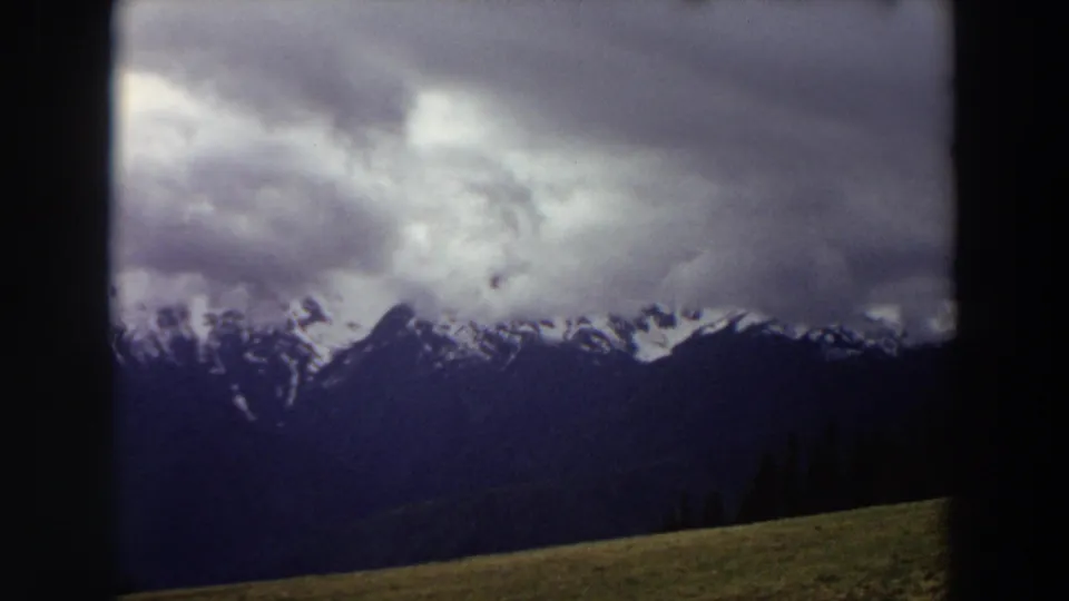 1964 Washington frame emphasizing heavy clouds over a dark snow-streaked mountain range, with a narrow grassy hillside in the foreground.