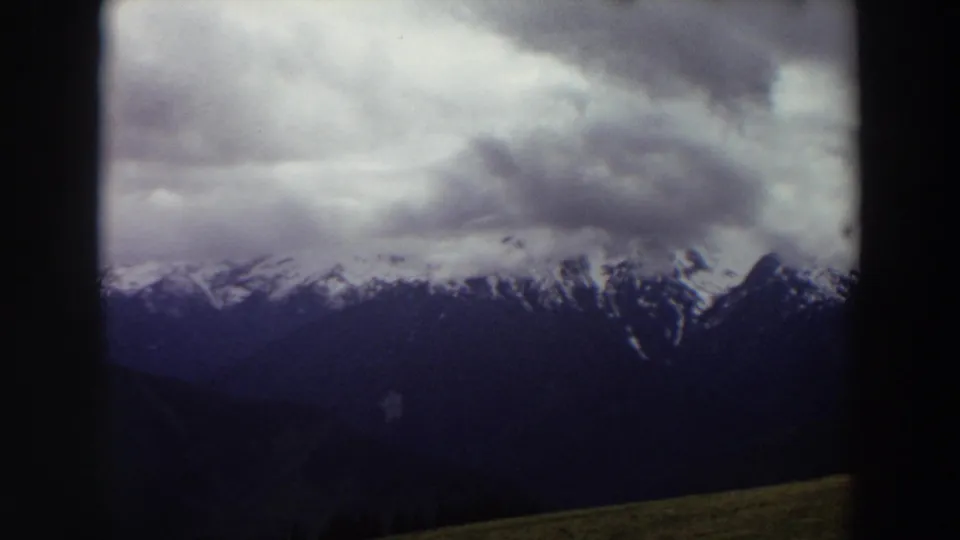 Washington 8mm frame from 1964 showing a distant snow-capped range beneath layered gray clouds and deep blue shadows.