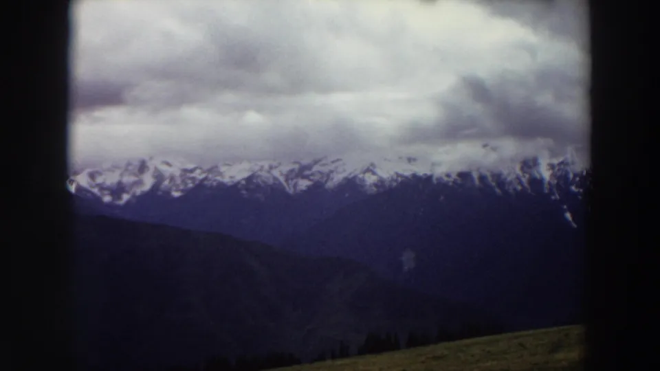 Late clip frame of a 1964 Washington mountain view, with snow-capped peaks under cloud layers and a grassy slope at the bottom.