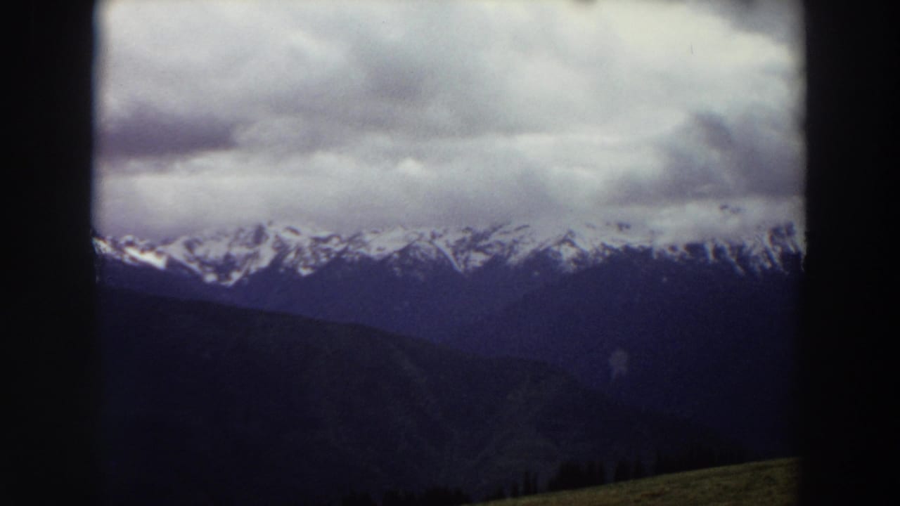 1964 Washington 8mm frame showing cloudy snow-capped mountains beyond dark ridges under a heavy sky.