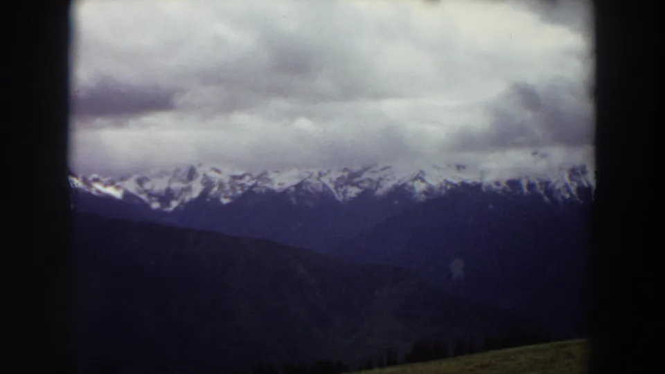 1964 Washington 8mm frame showing cloudy snow-capped mountains beyond dark ridges under a heavy sky.