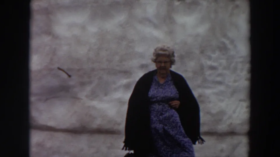 1964 Washington home-movie frame of an older woman waiting outdoors, her patterned dress shifting in the wind.