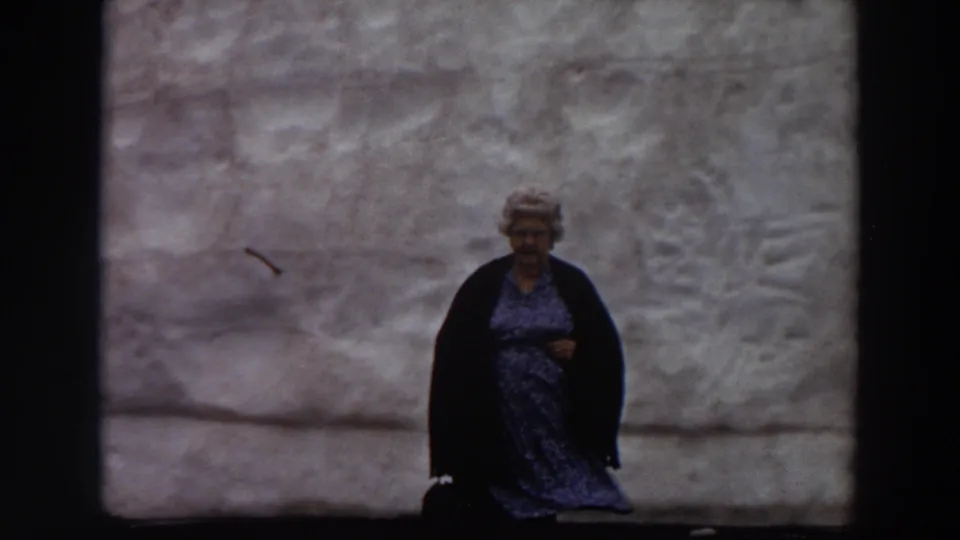 In 1964 Washington home-movie footage, a woman in a blue patterned dress stands near the center of a massive grey wall.