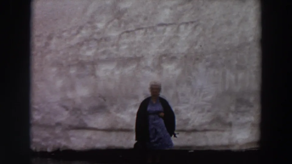The woman remains posed in front of the large grey wall, appearing smaller in the frame as the home-movie camera holds steady.