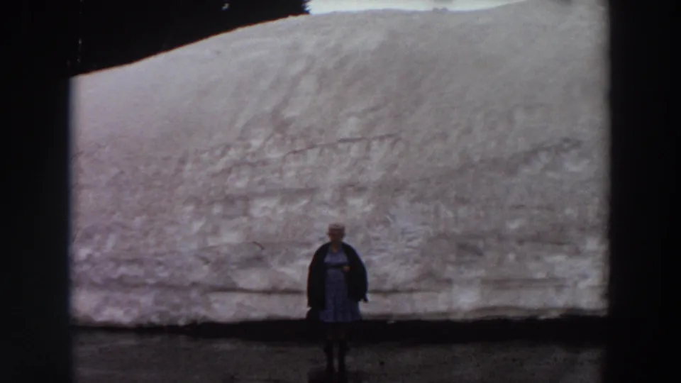 The final frame keeps the woman centered and small beneath the large grey wall, with muted 1960s 8mm color and visible frame edges.