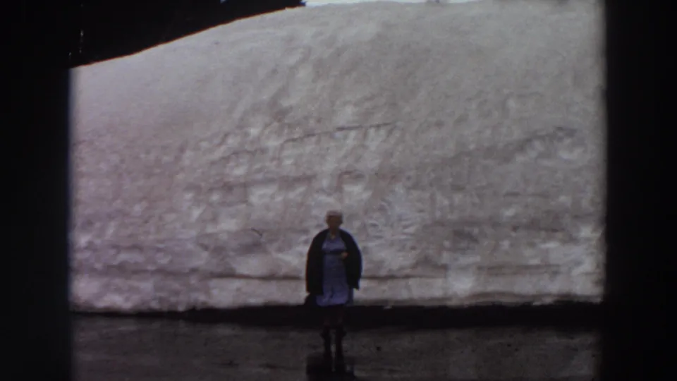 In 1964 Washington home-movie footage, a woman in a blue dress and dark cardigan poses before a massive snow wall.