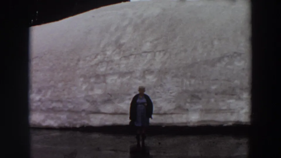 A woman stands centered against a broad snowbank or glacier-like surface in faded 1960s 8mm footage from Washington.