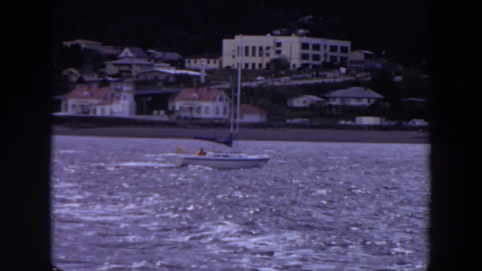 A sailboat passes along a Washington waterfront, with houses, larger buildings, and a dark hillside behind it.