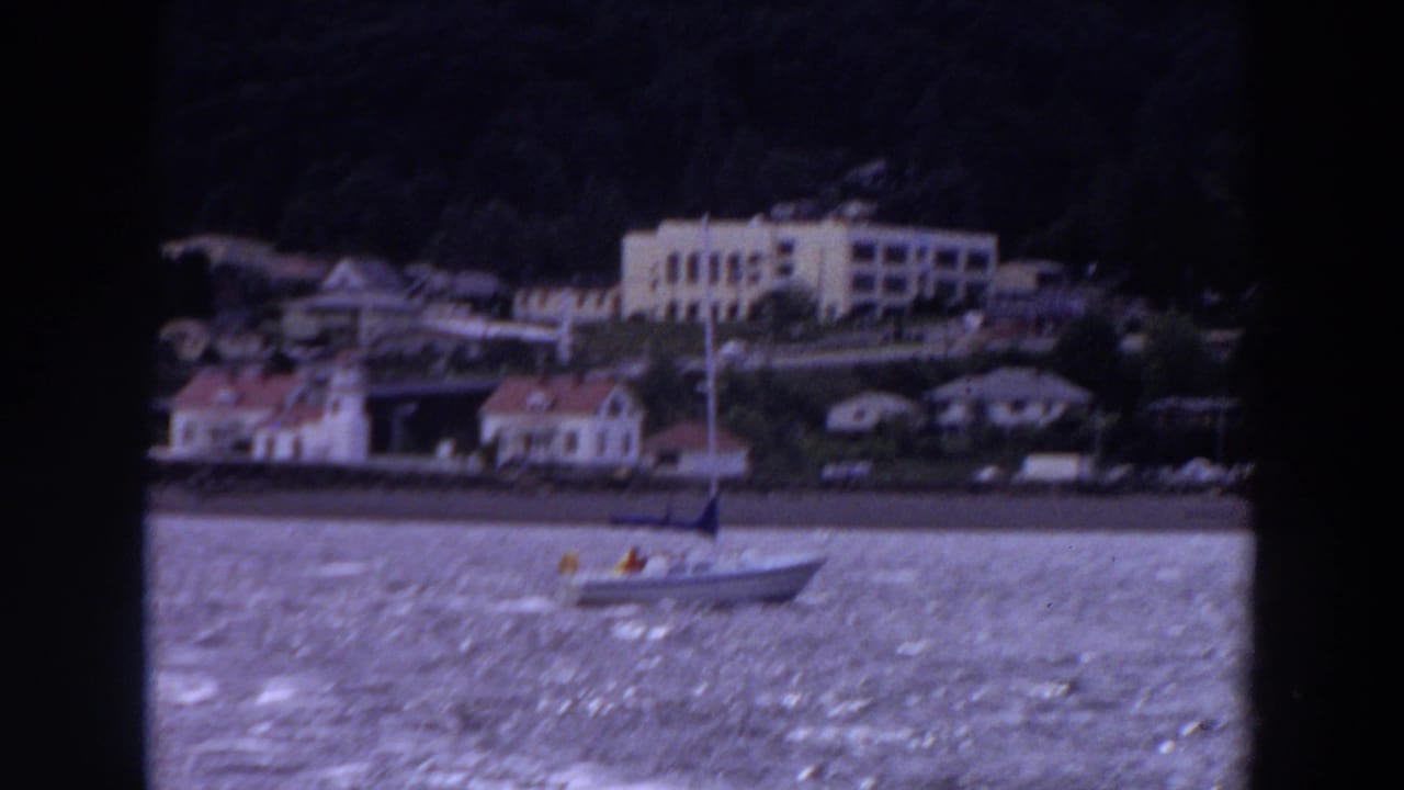 1964 Washington home-movie frame showing a small sailboat crossing rippled water below shoreline houses and a pale building on a wooded slope.