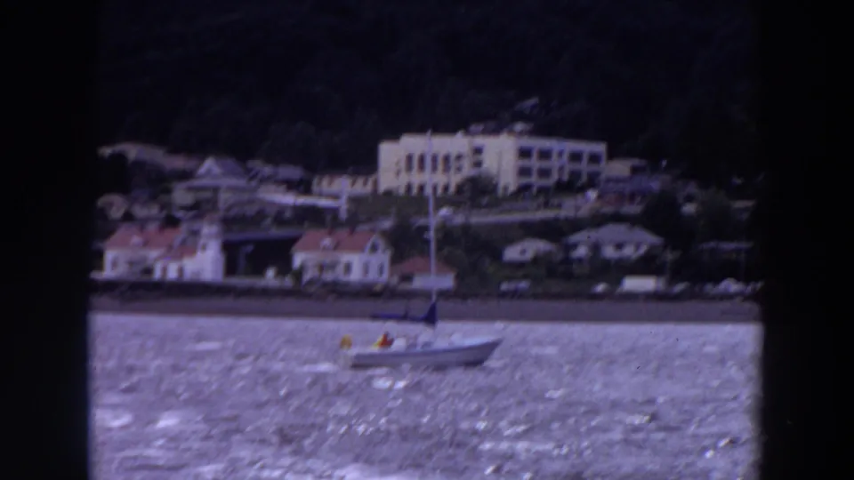 1964 Washington home-movie frame showing a small sailboat crossing rippled water below shoreline houses and a pale building on a wooded slope.