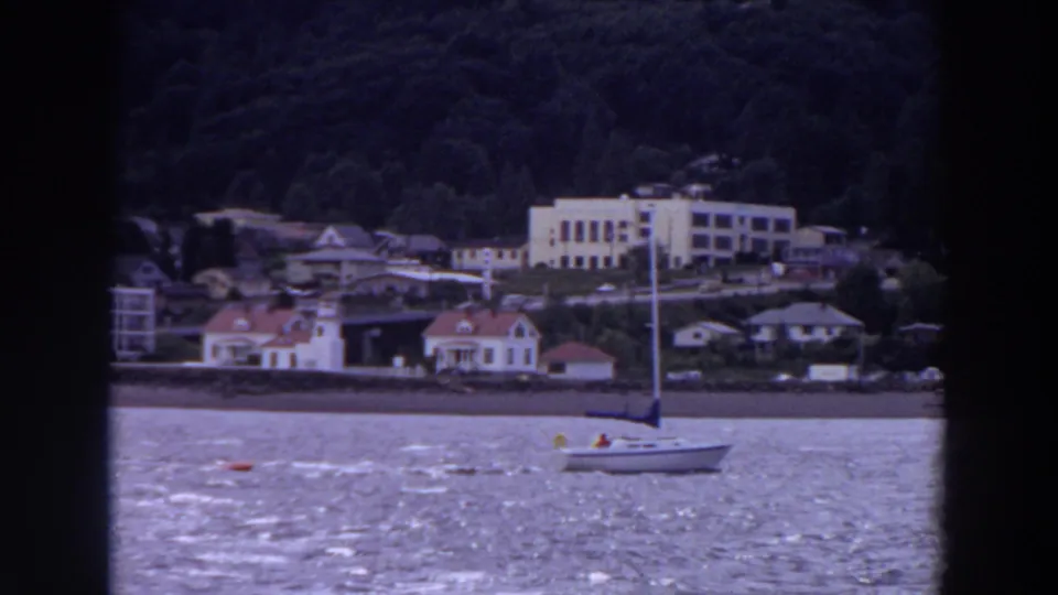 1964 Washington waterfront frame with a small sailboat moving across bright water in front of houses, trees, and a pale hillside building.