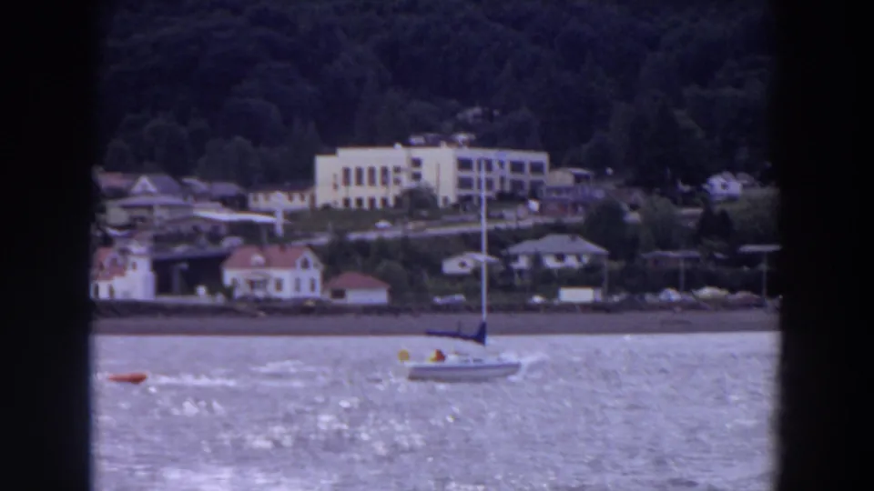1964 Washington 8mm frame showing a sailboat near a red buoy with waterfront houses and a pale hillside building in the background.