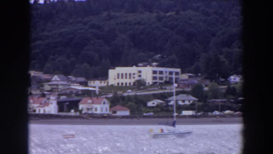 Late-frame view from 1964 Washington footage with a small sailboat at lower right and a settled wooded shoreline behind it.
